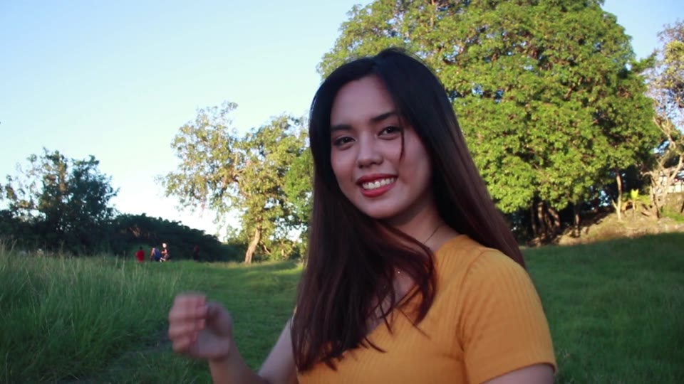 Original video frame: a woman with long dark hair raising her hand in a sunny park with trees and grass behind her
